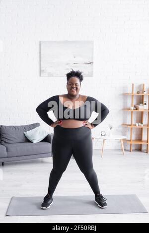 Volle Länge erfreut afroamerikanischen Übergewicht Frau in Sportbekleidung Zu Hause mit den Händen auf den Hüften stehen Stockfoto