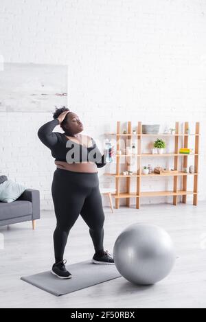 Müde afroamerikanische plus size Frau in Sportbekleidung halten Sport Flasche und Anpassung lockiges Haar Stockfoto