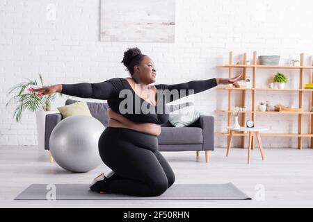 Happy african american plus size Frau in Sportswear mit ausgestreckt Hände trainieren im Wohnzimmer Stockfoto