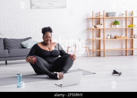 Happy african american plus size Frau in Sportswear Ausübung in der Nähe Laptop im Wohnzimmer Stockfoto