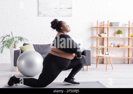 Seitenansicht der afroamerikanischen plus size Frau in Sportswear Ausfallschritte im Wohnzimmer Stockfoto