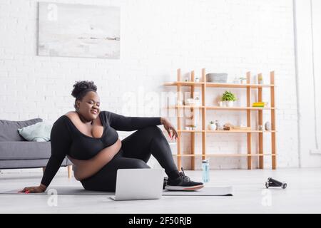 Lächelnd afroamerikanisch plus size Frau beobachten Online-Training auf Laptop, während Sie auf der Fitnessmatte sitzen Stockfoto