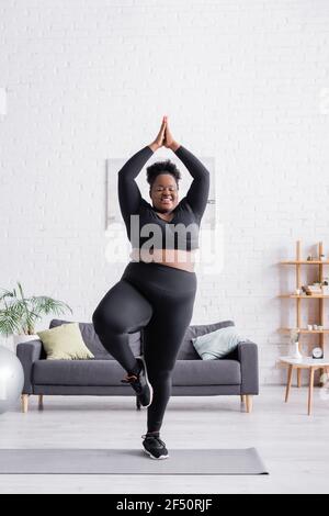 Happy african american plus size Frau in Sportswear stehend in Yoga-Pose im Wohnzimmer Stockfoto