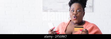 Schockiert afroamerikanische plus size Frau mit Kreditkarte und Smartphone, Banner Stockfoto
