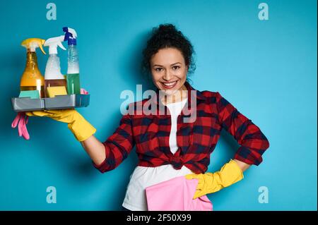 Lächelnde und glückliche lateinamerikanische Frau, die ihren Arm mit einem Tablett voller chemischer Reinigungsprodukte in der Hand hochhebt und das Ende der Hausarbeit genießt. Stockfoto