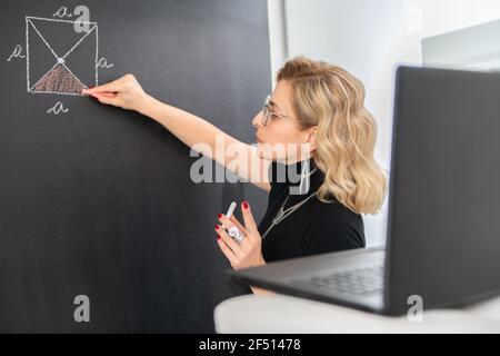 Junge Mathematiklehrerin erklärt Geometrie an der schwarzen Wand Stockfoto