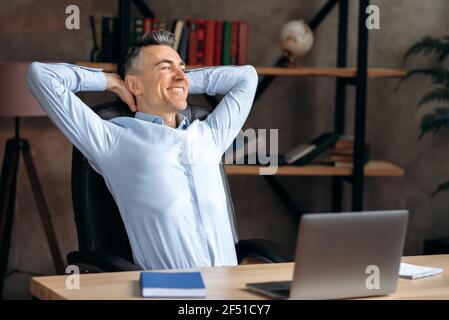 Zufriedener selbstbewusster kaukasischer Geschäftsmann, Rechtsanwalt oder Büroangestellter, entspannt am Arbeitsplatz und lächelt, sitzt in einem modernen Büro, ruht von der Arbeit, träumt von einem Urlaub, schaut weg Stockfoto