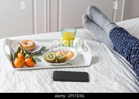 Horizontales Bild eines Tabletts mit Frühstück im Bett Neben einem schwarzen Handy und den Beinen einer Frau In blauen Pyjamas und grauen Socken auf einem Bett mit Ein Weiß Stockfoto