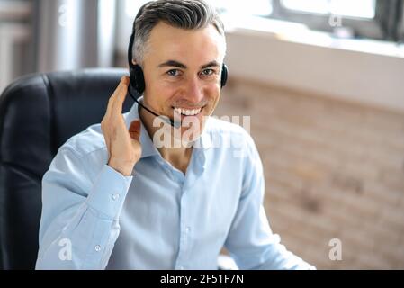 Portrait des erfolgreichen, selbstbewussten, gutaussehenden Geschäftsmanns, Managers oder Callcenter-Arbeiters, sitzt in einem modernen Büro, nutzt Kopfhörer für die Kommunikation mit dem Kunden, schaut auf die Kamera, freundliches Lächeln Stockfoto