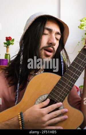 Junger langhaariger Mann spielt zu Hause akustische Gitarre und singt. Junger Bohemian. Trägt ein rosa Hemd mit einem Eimer Hut. Vertikales Bild Stockfoto