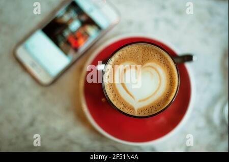 Nahaufnahme einer Kaffeetasse von oben mit einer Herzform aus Schaum und einem Smartphone auf einer Marmorplatte. Liebe Kaffee / Kaffeepause Konzept Stockfoto