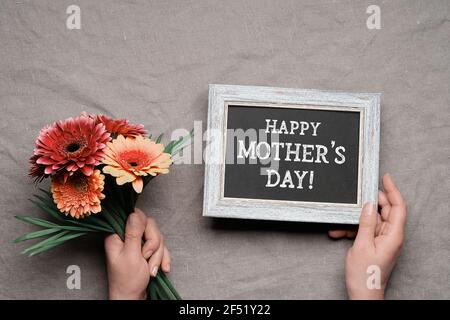 Frohes Muttertag, Kreidetext auf schwarzem Brett. Hände halten Tafel und Bund von orange, rot und lila Gerbera Blumen. Natürliche Erdfarben Stockfoto