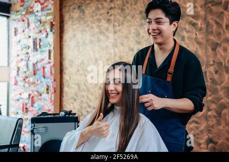 Portrait von glücklich und fröhlich junge weibliche Kunde zeigt thumbsup schild mit männlichen Friseur trägt Schürze Styling Haar Stockfoto