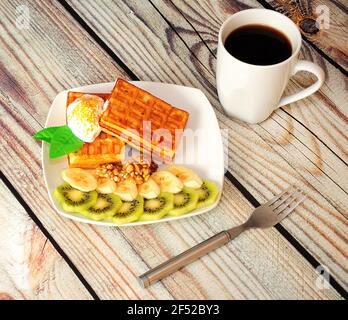 Köstliches Dessert, Wiener Waffeln mit Eis und Kiwi und Bananenscheiben auf einem Holztisch neben einem Kaffeebecher. Nahaufnahme. Stockfoto