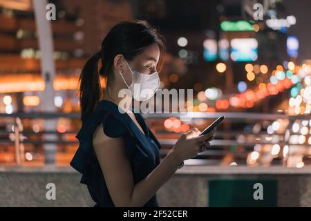 Seitenansicht der schönen jungen asiatischen glücklichen Frau mit covid-19 Gesicht Maske steht über Fußgängerbrücke Überprüfung Smartphone und Browsen durch Apps Beim Absuchen von E-Mails Stockfoto