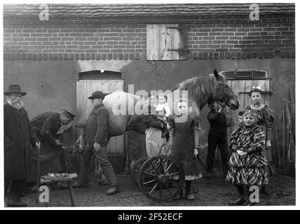 COSPA in Eilenburg / Deutschland: Gruppenbild (Familie Lübecks?) Vor dem Stallgebäude einer Pferdeernte. Im Hof links draußen ist ein älterer Mann mit Hut und Mantel, rechts draußen zwei Mädchen, die beide sowohl wohlhabende als die umliegenden Personen gekleidet sind. Gruppenbildung mit Bauern, einer Mutter mit Kind und dem Hufschmied, der gerade ein Pferd schiebt Stockfoto