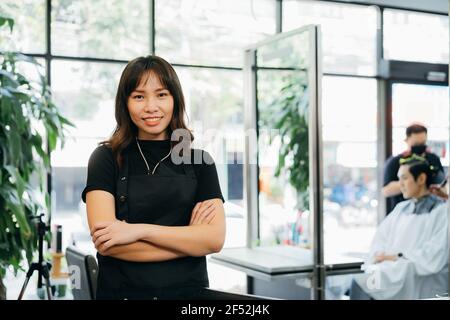 Porträt von glücklich erfolgreichen jungen asiatischen Salon Besitzer stehend tragen Schürze mit gefalteten Händen lächelnd beim Blick auf die Kamera Stockfoto