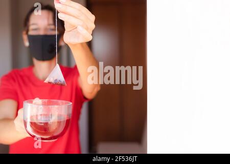 Frau mit rotem Kleid ist einweichen Teebeutel auf Kristallbecher, Vorbereitung heißen Tee. Teebeutel auf Tasse tauchen. Frau während der Quarantäne. Es gibt eine weiße Wand für Stockfoto