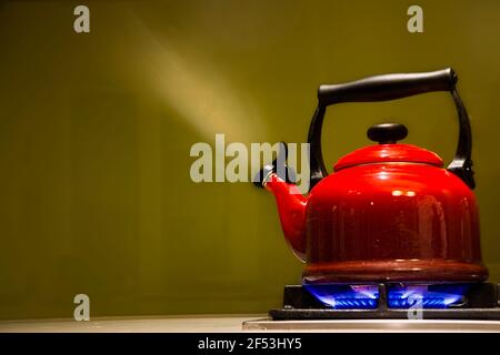 Dampfender hellroter Wasserkocher auf Gasherd mit blauen Flammen vor grünem Hintergrund in moderner Küche. Stockfoto