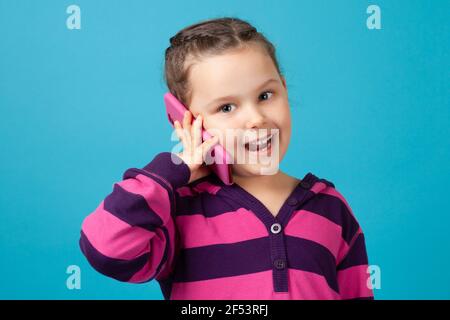 Glückliches Mädchen mit Zöpfen hält ein Telefon in der Nähe ihres Ohres und macht einen Anruf, Kommunikation mit Verwandten oder Freunden, isoliert auf einem blauen Hintergrund Stockfoto