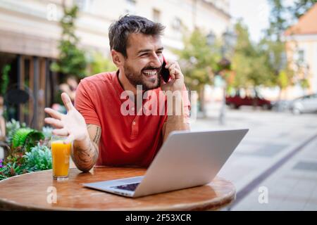 Smliling man spricht auf einem Telefon, während er im Café vor einem Laptop sitzt Stockfoto