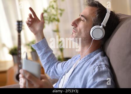 Detail des Mannes sitzt auf dem Sofa zu Hause hören Musik von einem Mobiltelefon mit weißen Kopfhörern Eintritt in ein warmes Sonnenlicht durch Th Stockfoto