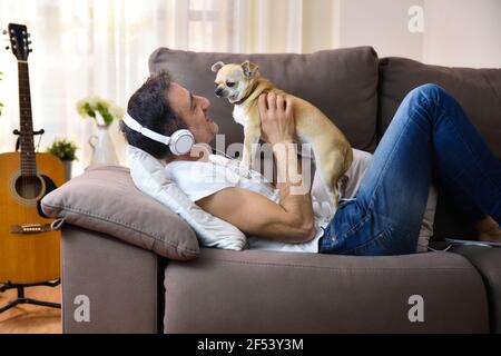 Mann, der mit seinem Hund spielt und liebevoll auf sein Gesicht schaut Liegen auf dem Sofa zu Hause Musik hören mit Weiße Kopfhörer Stockfoto