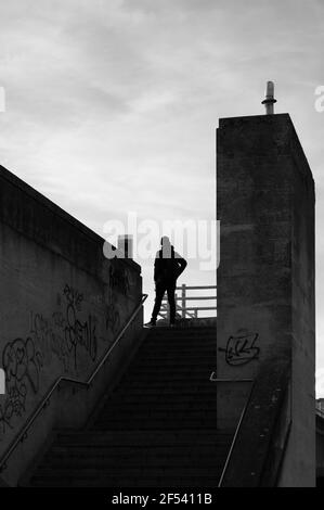 Eine schwarz-weiße Silhouette eines einsamen Mannes, der Hand auf Hüfte oben auf einer äußeren Betontreppe mit Graffiti an den Wänden steht Stockfoto