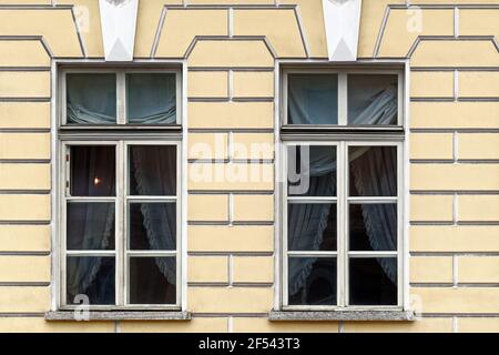 Zwei rechteckige Fenster mit alten Holzrahmen und Vorhängen an einer gelben Stuckwand. Aus den Fenstern der St. Petersburg-Serie. Stockfoto