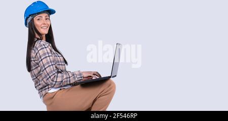 Banner, Langformat. Kaukasische Frau Handyman in Hut und mit tragbaren Laptop sitzt auf grauem Hintergrund mit Seitenfläche für Text oder Werbung. Stockfoto