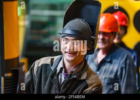 Ekibastuz, Region Pavlodar, Kasachstan: Eisenbahnkraftwagenbau. Junger Schweißer in Schutzmaske lächelnd und posiert. Andere Personen verschwommen. Stockfoto