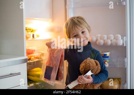 Cute Kleinkind blonde Kind, Öffnen des Kühlschranks doo Rand aus geräuchertem Fleisch, Prosciutto, gesunde Lebensmittel im Kühlschrank Stockfoto