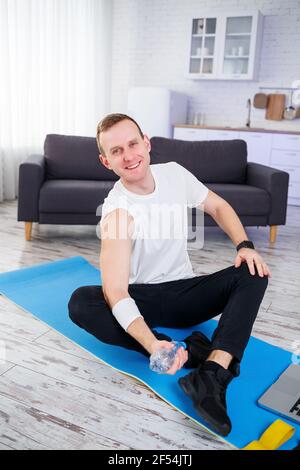 Hübscher junger Mann, der auf dem Boden sitzt und die Flasche hält Des Wassers während des Online-Trainings Stockfoto