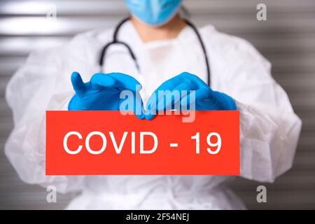 Nahaufnahme des nicht erkennbaren Gesundheitspersonals, das chirurgische Masken und Schutzhandschuhe trägt, die ein Schild aus Pappe mit dem Text Covid-19 halten. Stockfoto