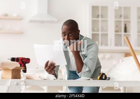 Probleme bei der Montage neuer Möbel. Schockiert afroamerikanischen Mann lesen Anweisungen zu Hause Stockfoto