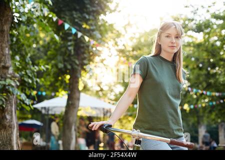 Mädchen auf dem Fahrrad. Porträt einer jungen Frau, die auf einem Fahrrad sitzt und verwirrt auf die Kamera schaut und in einem Stadtpark oder einem grünen Faire lächelt. Hipster Stockfoto