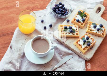 Waffeln mit Sahne und Heidelbeeren, eine Tasse Kaffee und Orangensaft auf dem Tisch auf einer Stoffserviette. Draufsicht. Stockfoto