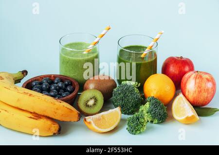 Zwei verschiedene grüne Smoothies in Gläsern mit Strohhalmen, Banane, Äpfel, Brokkoli, Kiwi, Zitrone und Heidelbeeren in einer Schüssel. Gesunde Zutaten, weißer Rücken Stockfoto