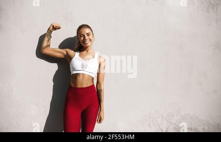 Porträt der jungen Fitness Frau, die Muskeln und lächeln. Kaukasische weibliche Modell in Sportbekleidung zeigt Bizeps, im Freien in der Nähe Betonwand stehen Stockfoto