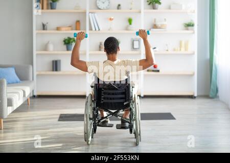 Rückansicht des behinderten schwarzen Kerls im Rollstuhl, der mit Hanteln zu Hause trainiert. Sport- und Behindertenkonzept Stockfoto