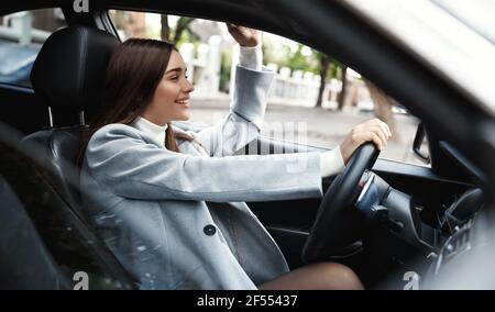 Unternehmen. Glückliche busienswoman Auto fahren und winken Hand an Freund. Elegante weibliche Fahrerin, die sich auf die Konferenz begibt und hallo sagt Stockfoto