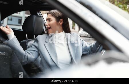 Geschäftsfrau, die Auto fährt, ärgerlich hinter sich schaut und sich beschwert. Weibliche Exekutive in Eile stecken im Verkehr, streiten mit einem anderen Fahrer Stockfoto