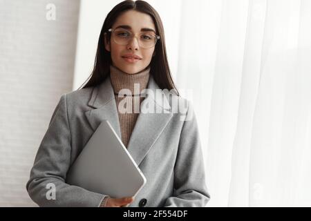Junge Geschäftsfrau in Brille und grauem Mantel, die im Büro steht, Laptop hält und auf die Kamera schaut Stockfoto