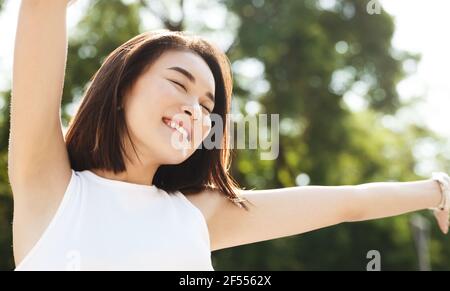 Nahaufnahme einer asiatischen Frau, die sich die Hände hochstreckt und lächelt, im Park läuft und unbeschwert und glücklich aussieht. Modernes Mädchen atmen frische Luft auf einem Spaziergang, schauen Stockfoto