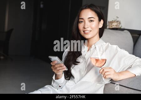 Nahaufnahme einer eleganten jungen asiatischen Frau, die sich zu Hause entspannt, auf dem Handy Online-Shopping-Websites durchstöbert und ein Glas Wein genießt. Weibchen ruht auf Stockfoto