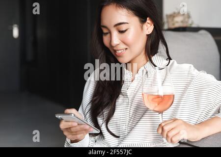 Nahaufnahme einer eleganten jungen asiatischen Frau, die sich zu Hause entspannt, auf dem Handy Online-Shopping-Websites durchstöbert und ein Glas Wein genießt. Weibchen ruht auf Stockfoto