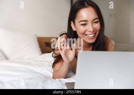 Fröhliche attraktive asiatische Mädchen im Bett mit Laptop liegen und winkende Hand an der Computer-Kamera, haben Videoanruf mit Freunden, während zu Hause bleiben Stockfoto