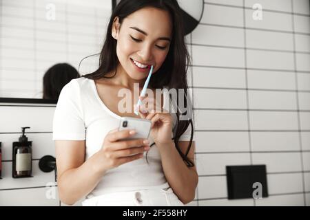 Lifestyle. Schöne asiatische Frau Zähneputzen und lesen Nachricht am Telefon aus dem Badezimmer. Mädchen mit Smartphone mit Zahnbürste, überprüfen sozialen Stockfoto