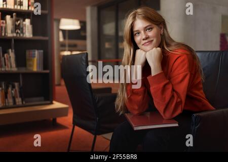 Portrait von jungen attraktiven Rotschopf weibliche Studentin, sitzen Sessel auf Armen und lächelnd, Kamera suchen, diskutieren Roman mit bookclub in Stockfoto