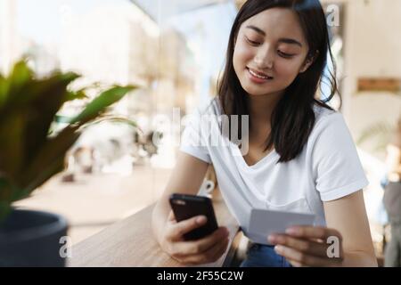Nahaufnahme einer glücklichen asiatischen Frau, die mit Kreditkarte und Smartphone bezahlt, während sie im Café neben dem Fenster sitzt und lächelt, während sie Geld sendet Stockfoto